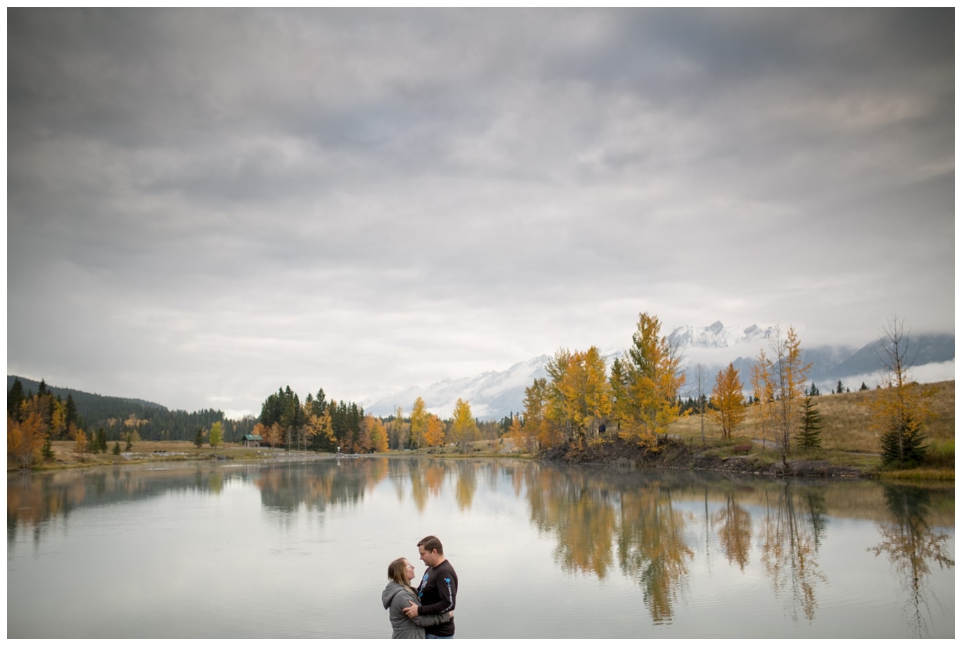 An engagement picture with the couple framed in a lake with mountains in the background, fall trees with bright yellow leaves, and a dramatic sky