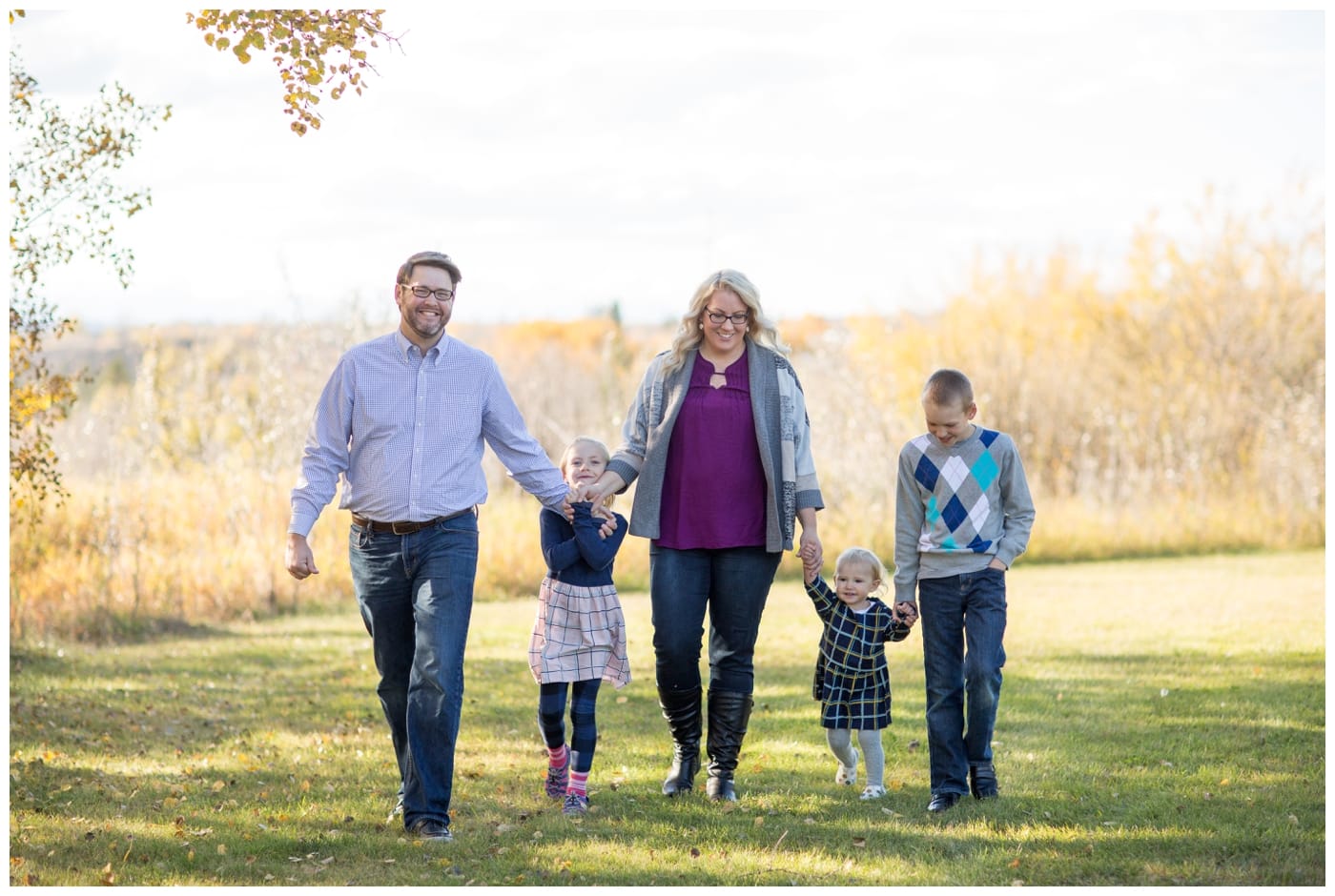 A family of five walk hand in hand along a field of grass while all smiling and laughing