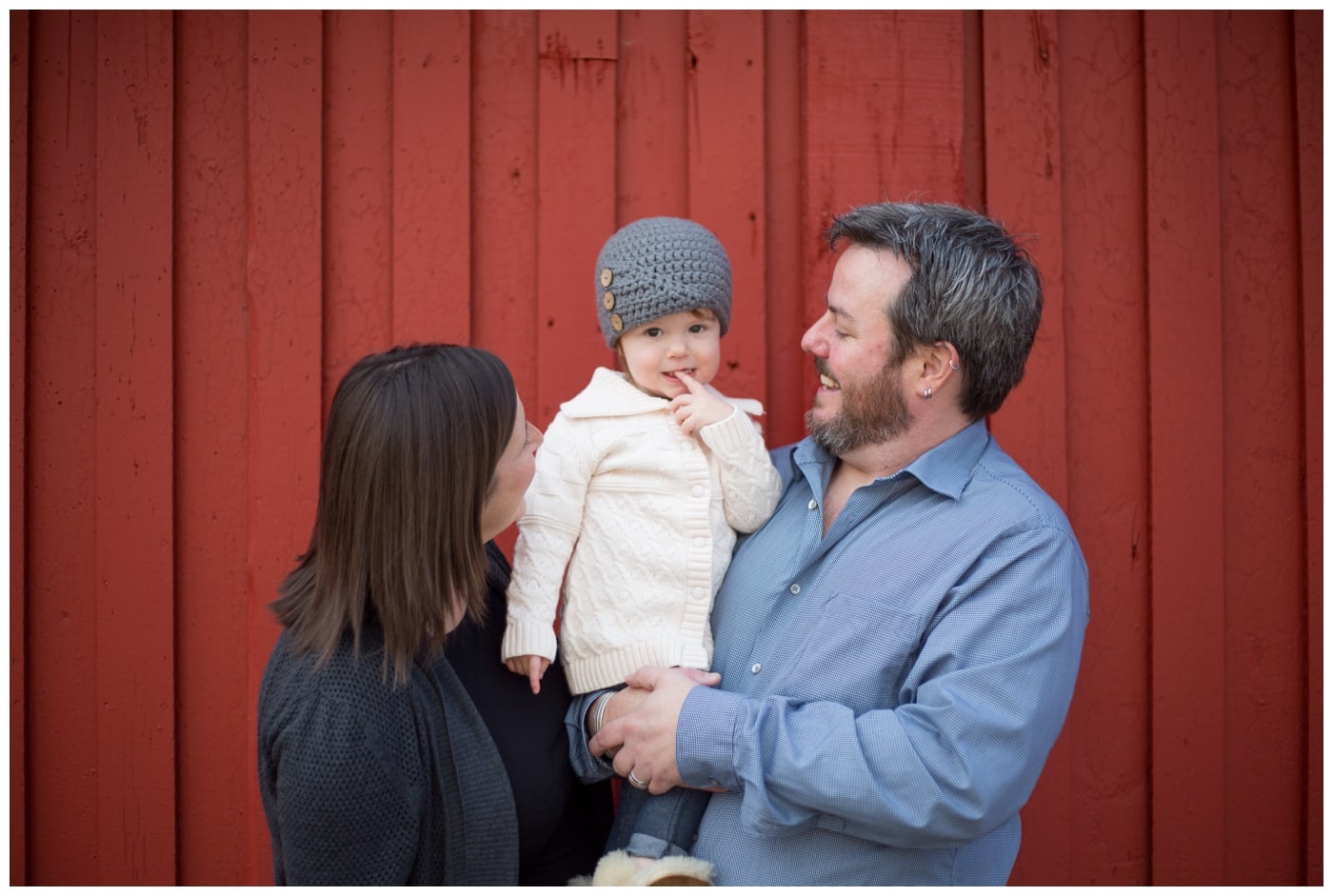 A young girls smiles at the camera while being held in her mom and dad's arms with a bright red wooden background