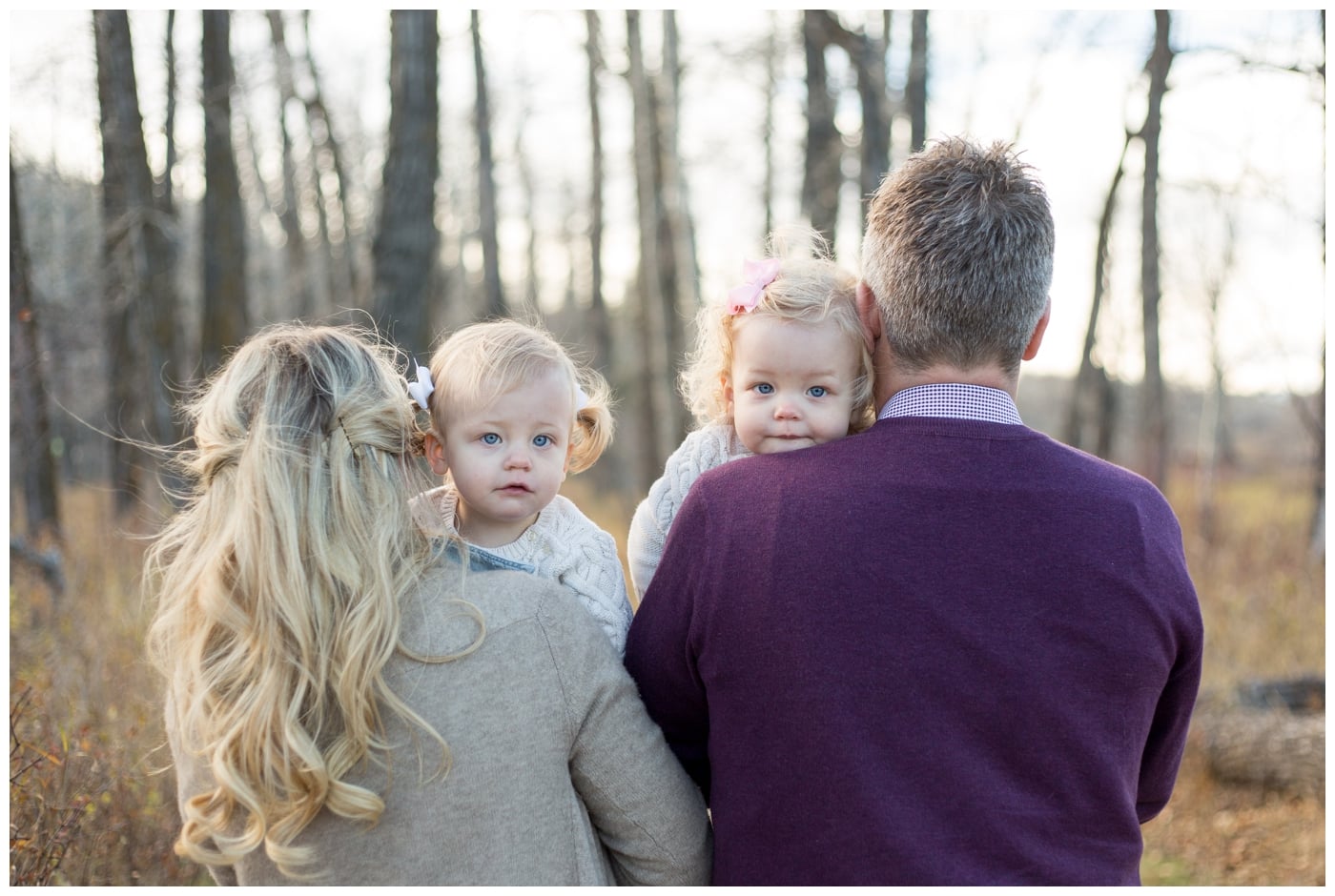 Two identical twin girls peek over their parents shoulders at the camera