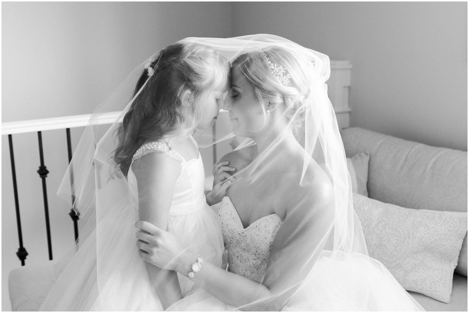 A black and white photo of a bride and her daughter cloaked in her veil as they sit on a couch