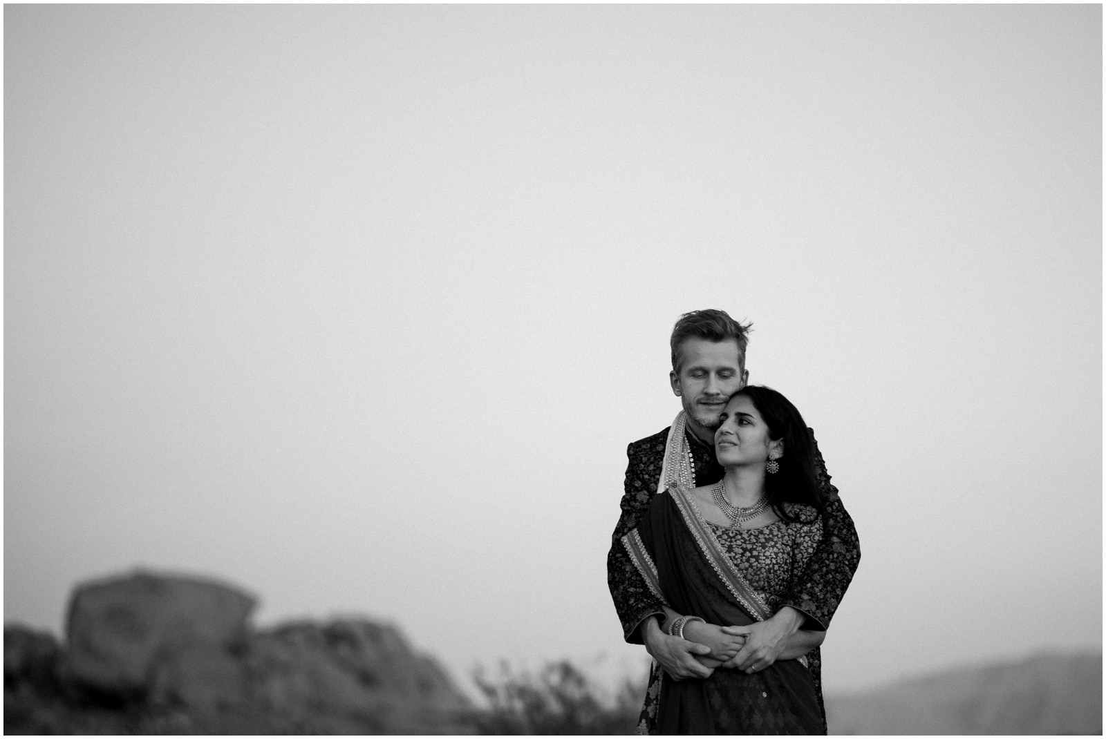 A black and white image of a bride and groom wearing traditional Indian dress embrasing each other in the last hour of sunset