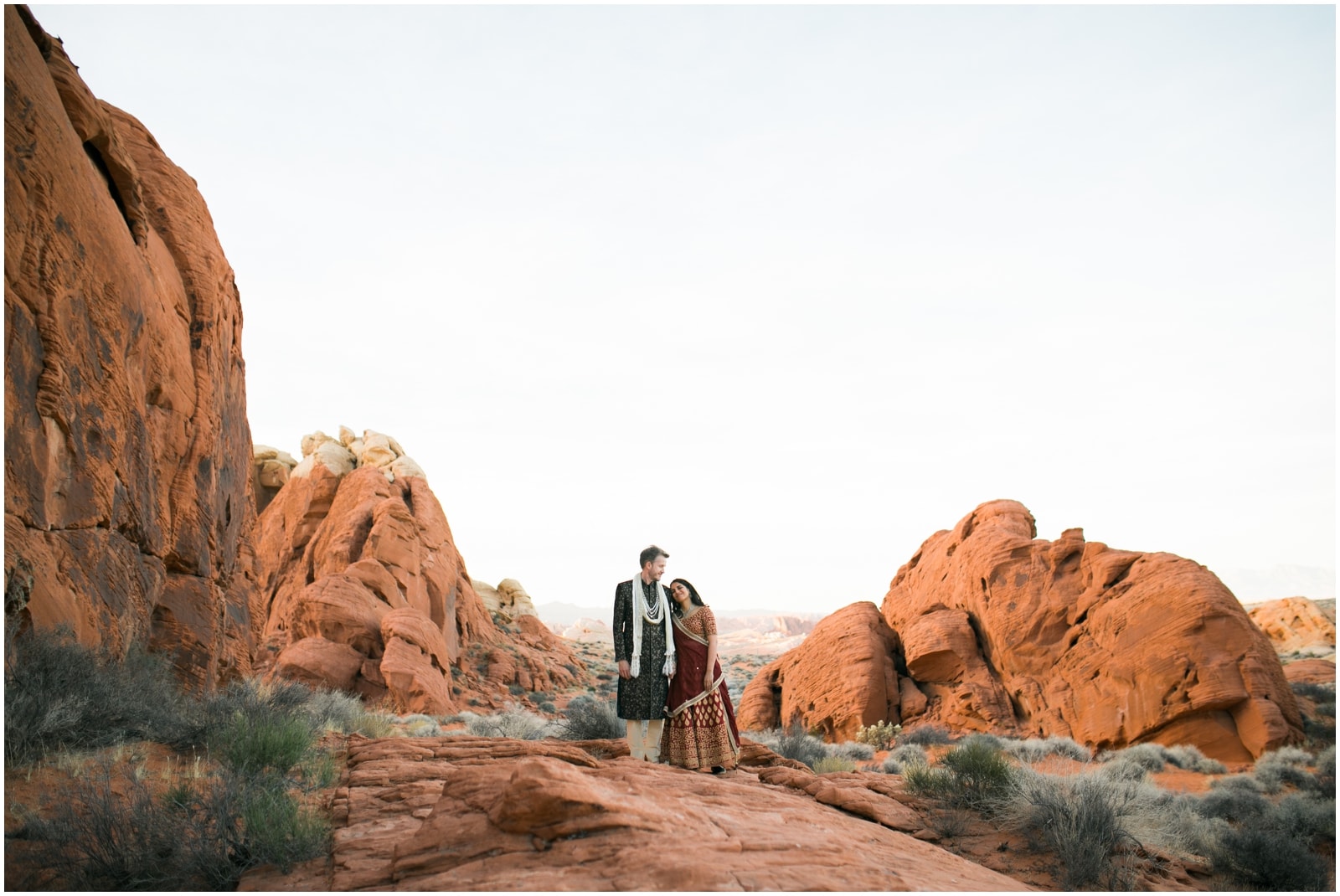 A bride rests her head on her groom's shoulder as they are surrounded by bright red rocks in the Valley of Fire Nevada