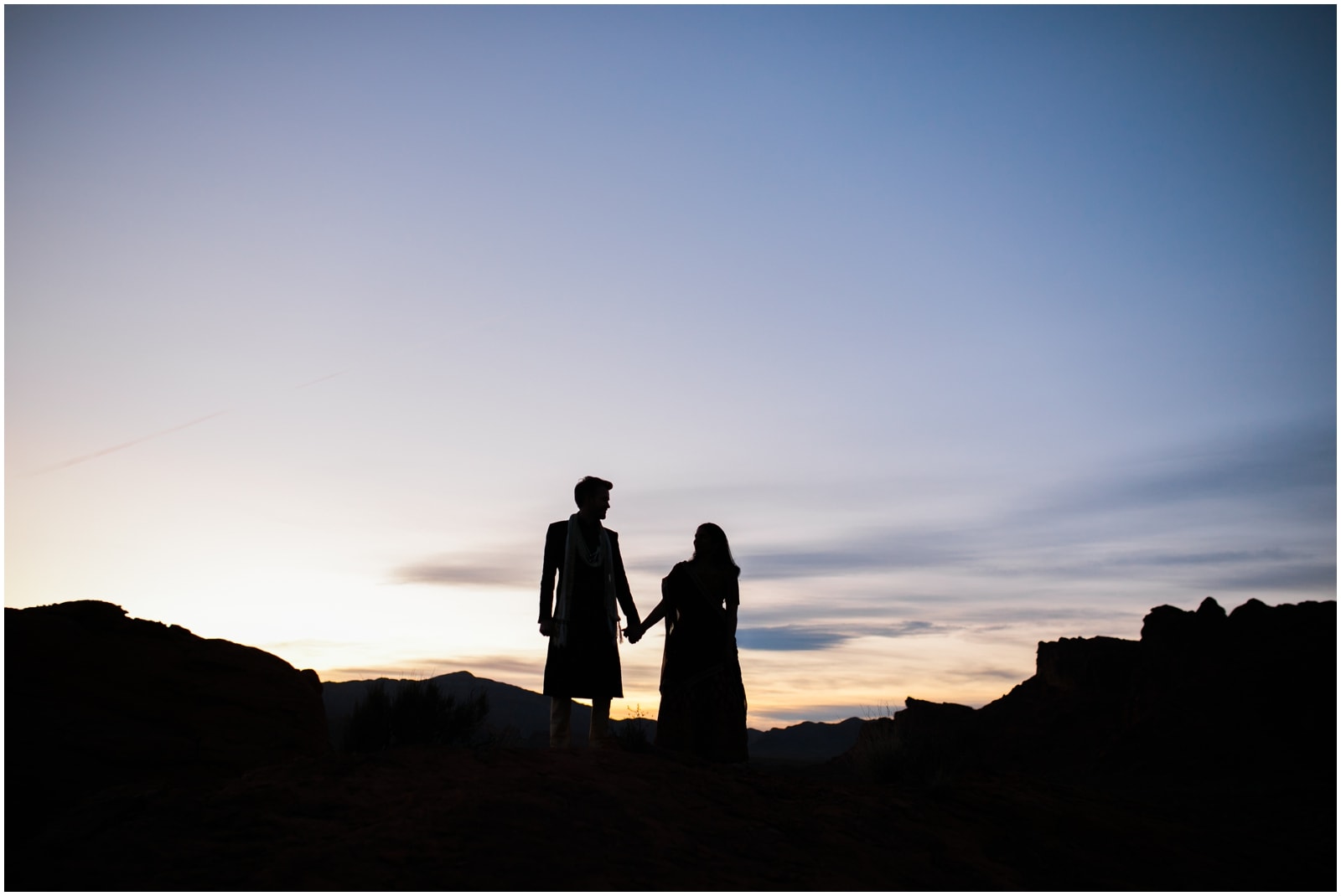 A bride and groom in traditional Indian dress are silhouetted holding hands against a setting sky