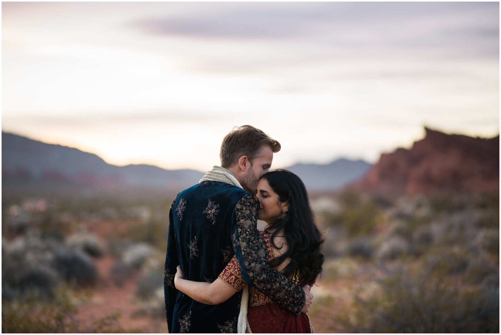 A bride and groom in traditional indian dress face away from the camera and snuggle their forheads together as they watch the sunset in the Nevada desert