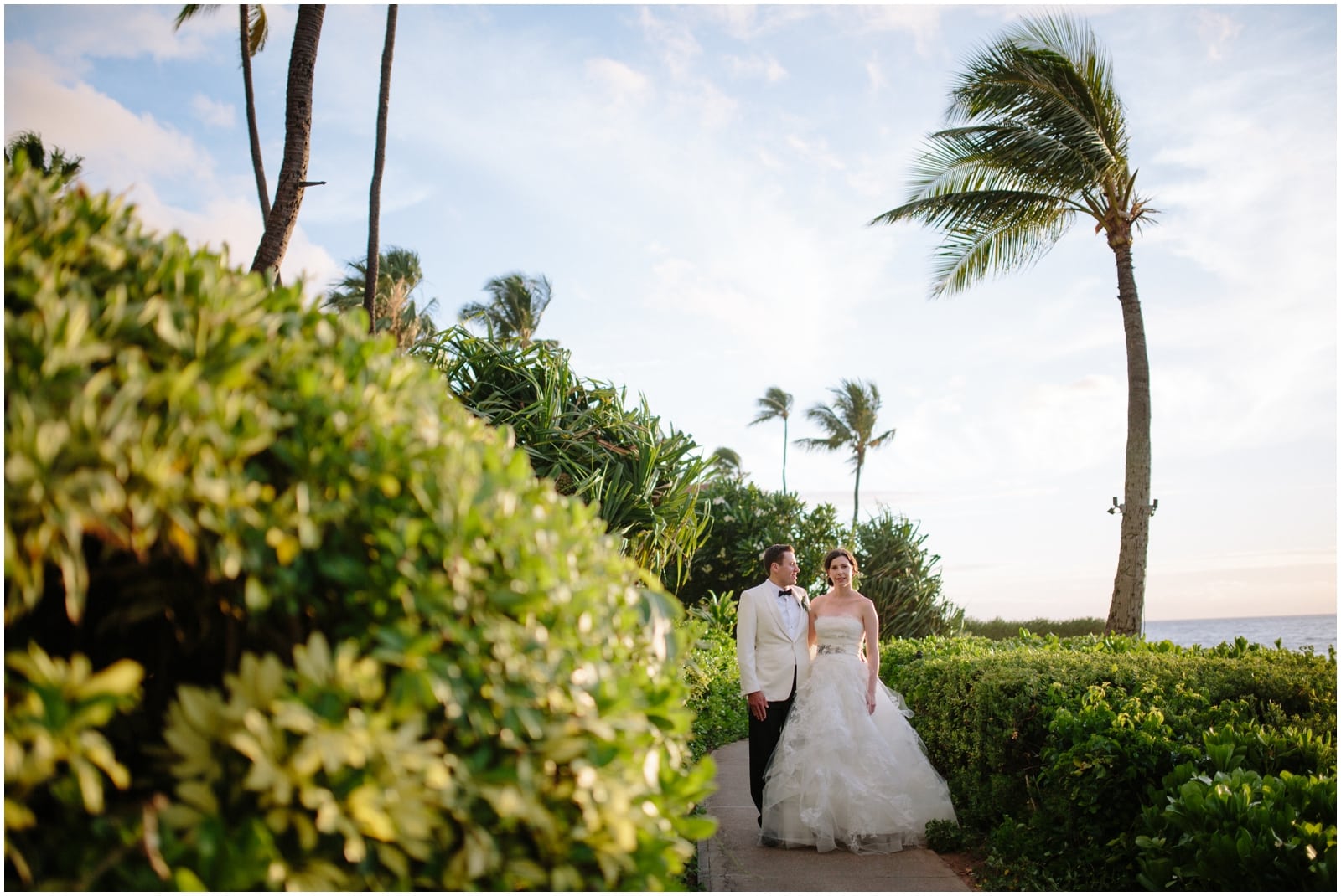 A bride and groom in Maui stay on a path beside palm trees and the ocean