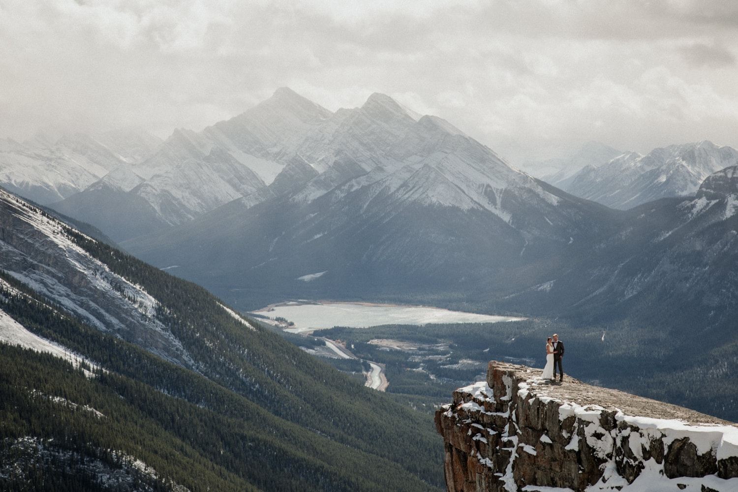 A couple in wedding attire stands atop a mountain in Banff National Park with epic mountain views and clouds in the background