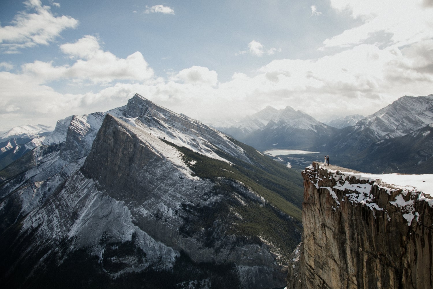 A couple in wedding attire stands atop a mountain in Banff National Park with epic mountain views and clouds and a lake in the background