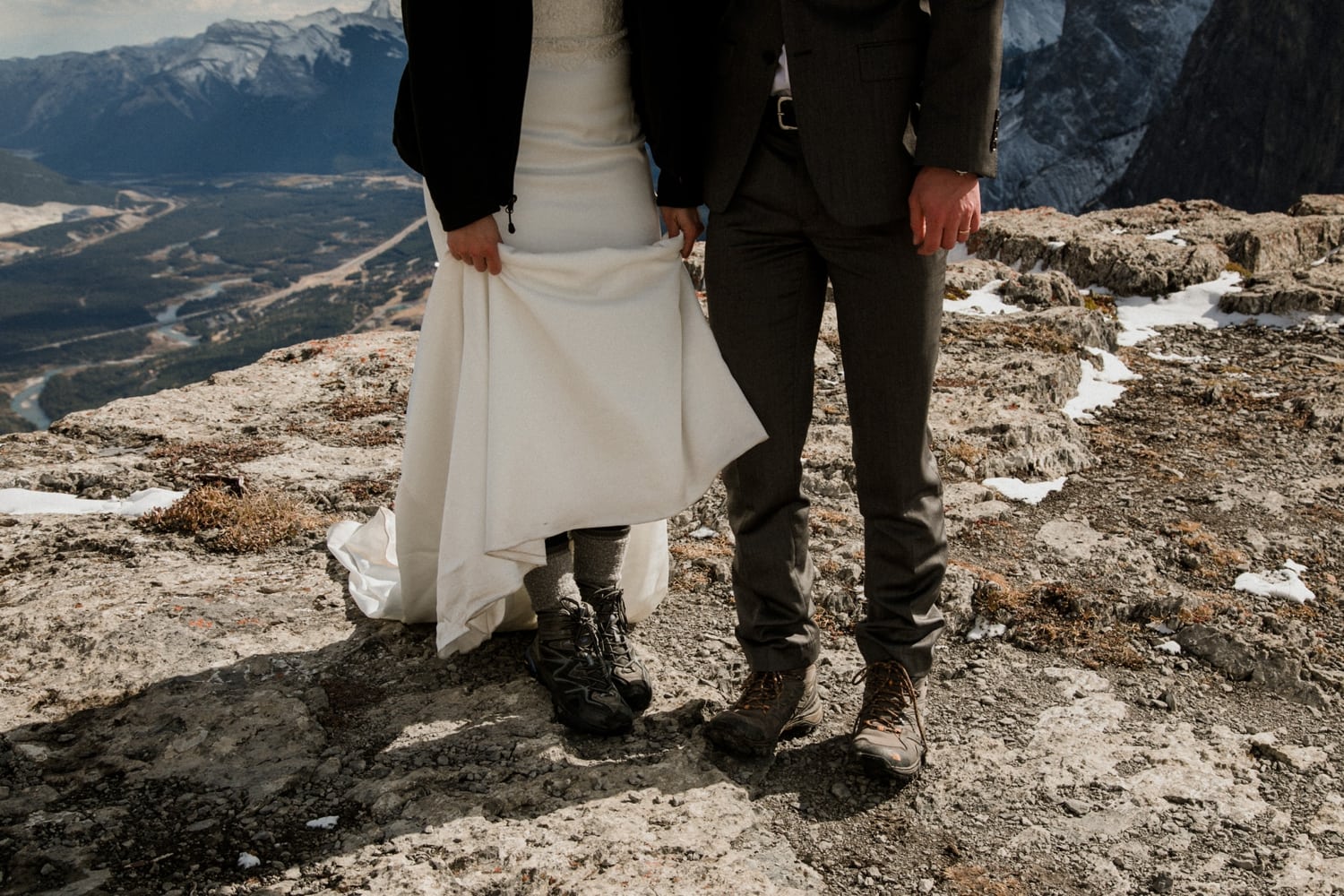 A couple standing on top of a mountain show off their hiking boots underneath their wedding attire