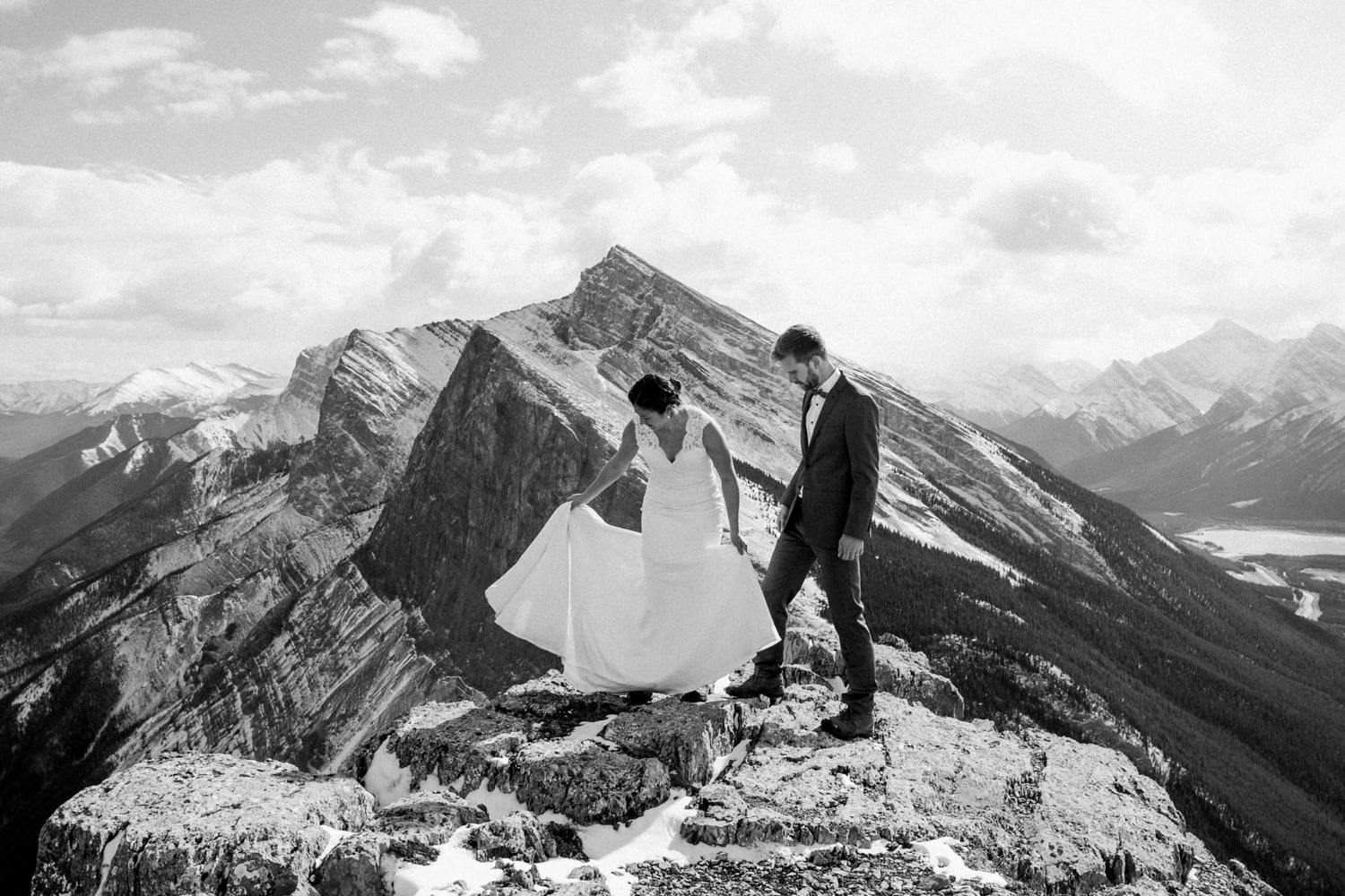 A black and white photo of a couple in wedding attire standing atop a mountain in Banff National Park with epic mountain views and the woman is adjusting her wedding dress