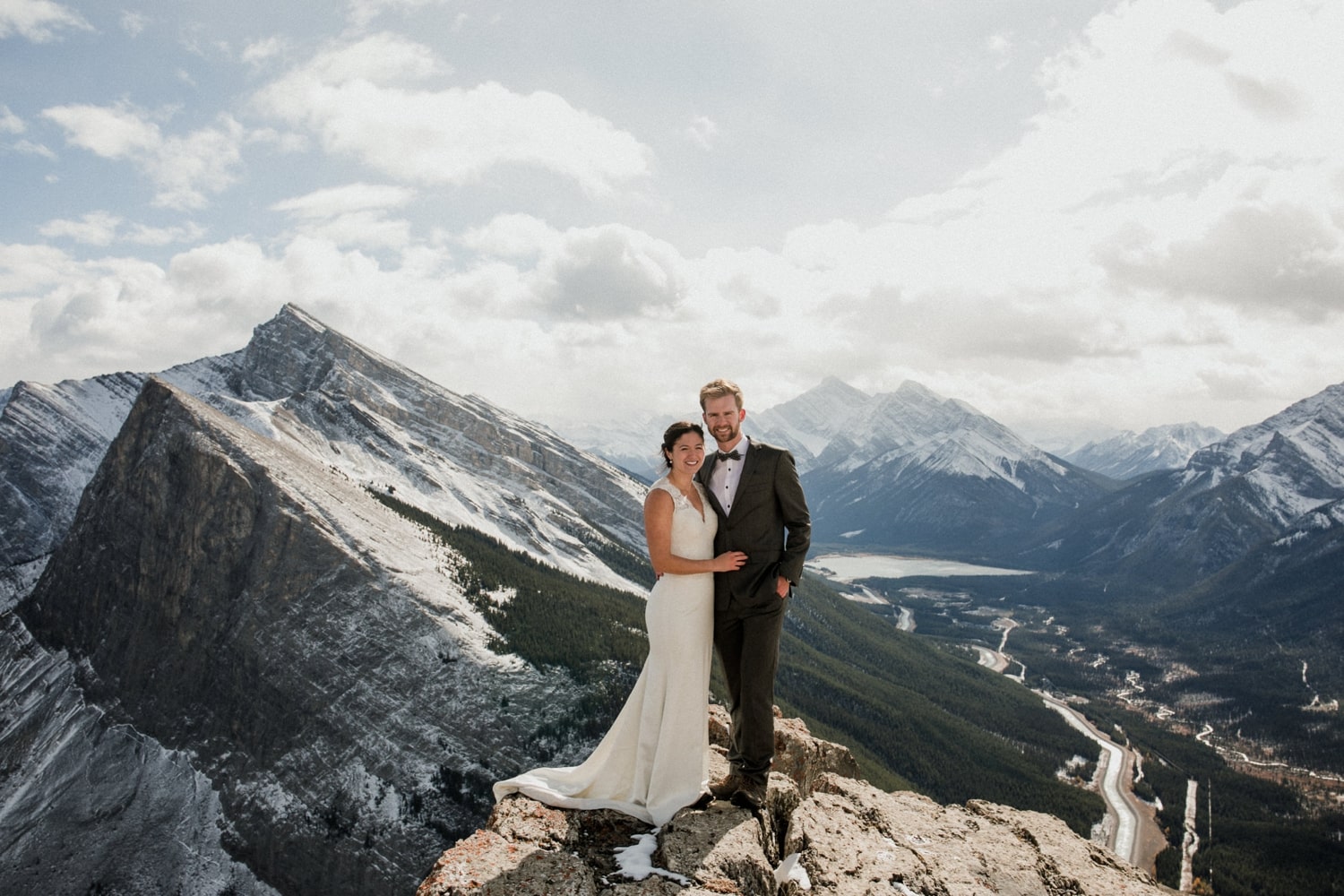 A couple in wedding attire stands atop a mountain in Banff National Park with epic mountain views and clouds in the background