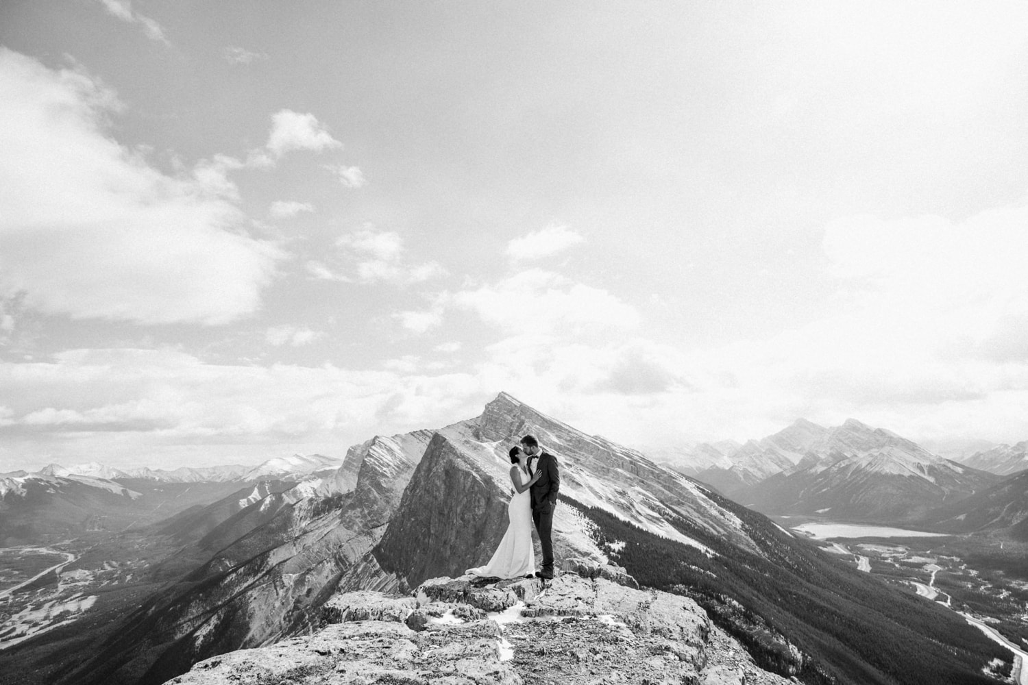 A black and white photo of a couple embrassing in a kiss on top of a mountain with sweeping mountainviews and clouds in the background