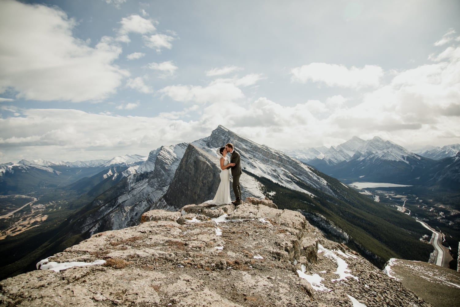 A photo of a couple embrassing in a kiss on top of a mountain with sweeping mountainviews and clouds in the background