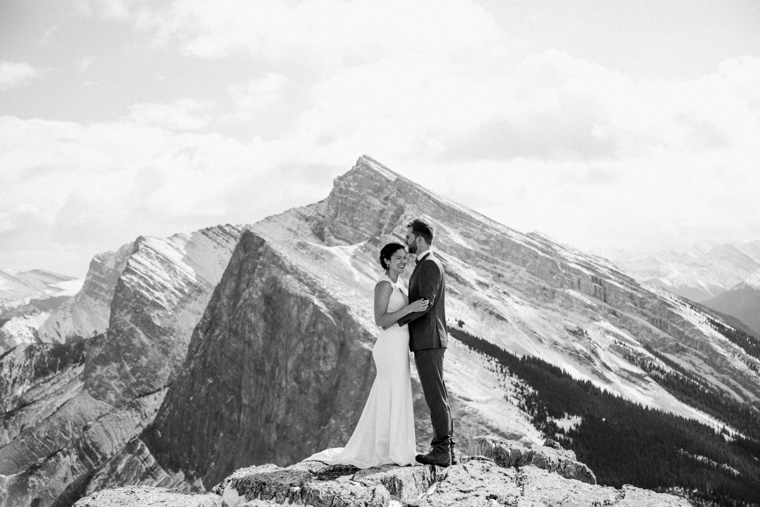 A black and white photo of a couple laughing as they stand on top of a mountain with sweeping mountainviews and clouds in the background