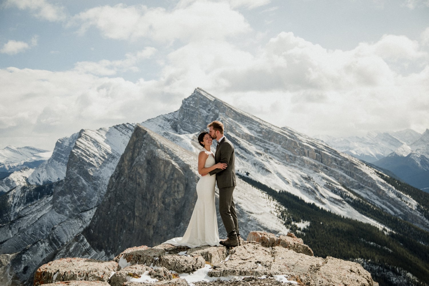 A photo of man kissing his bride on the cheek on top of a mountain with sweeping mountainviews and clouds in the background