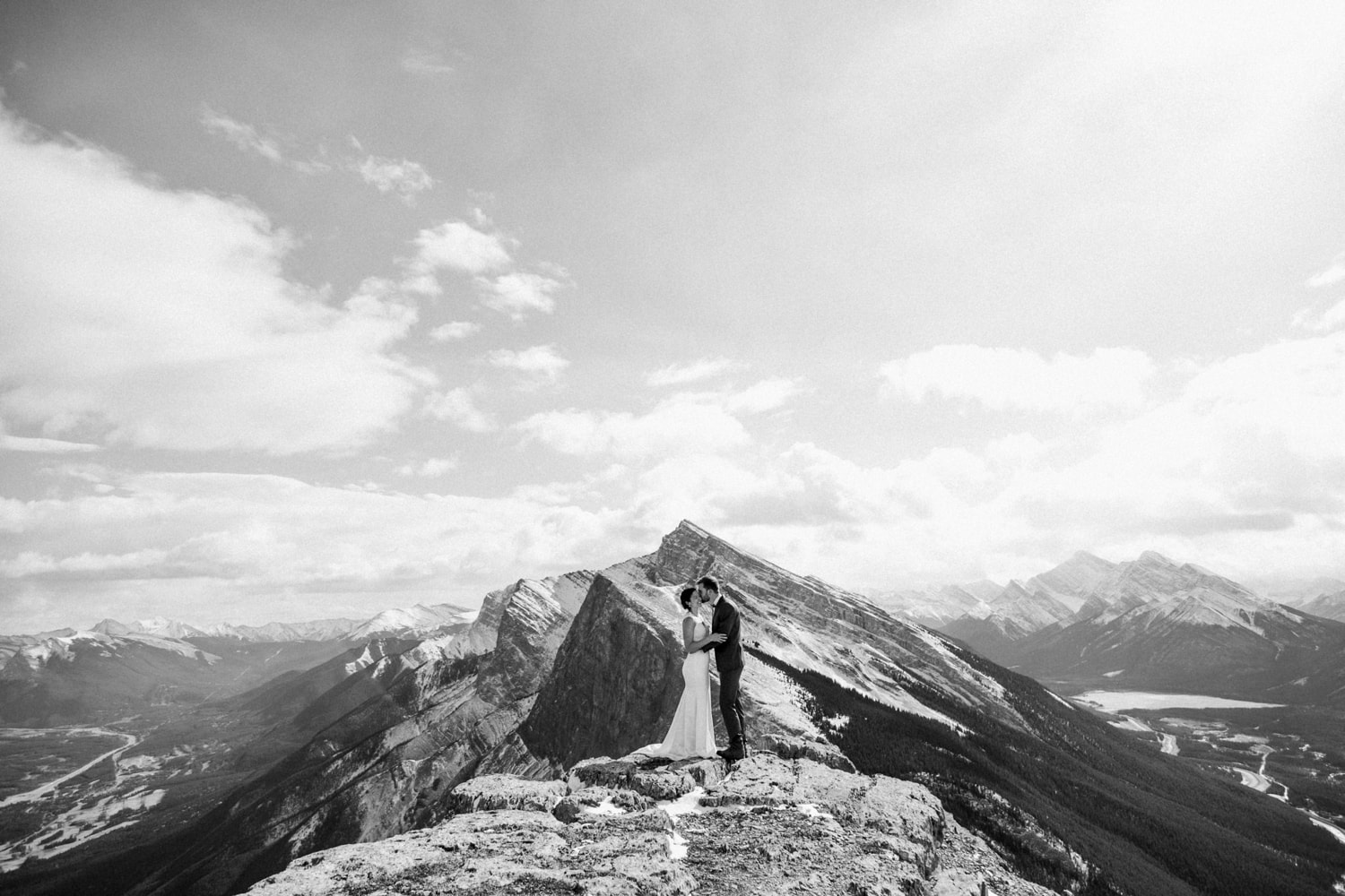 Black and white photo of a couple standing on a mountain with mountains framing behind them and clouds in the sky