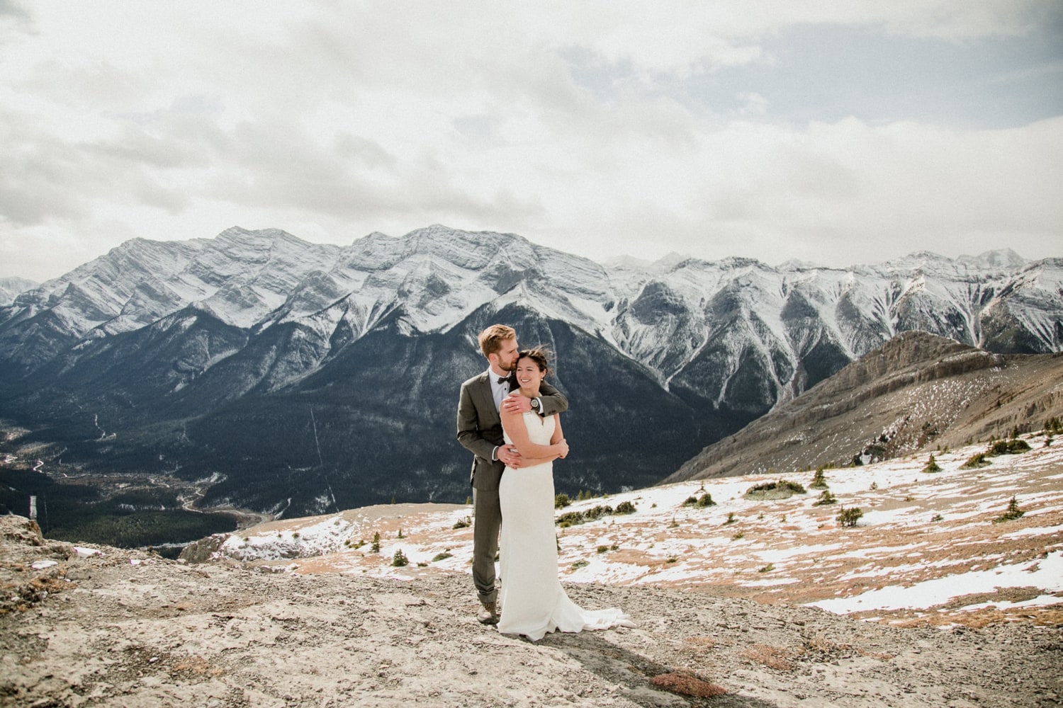 A photo of a couple hugging on top of a mountain with sweeping mountainviews and clouds in the backgroundv