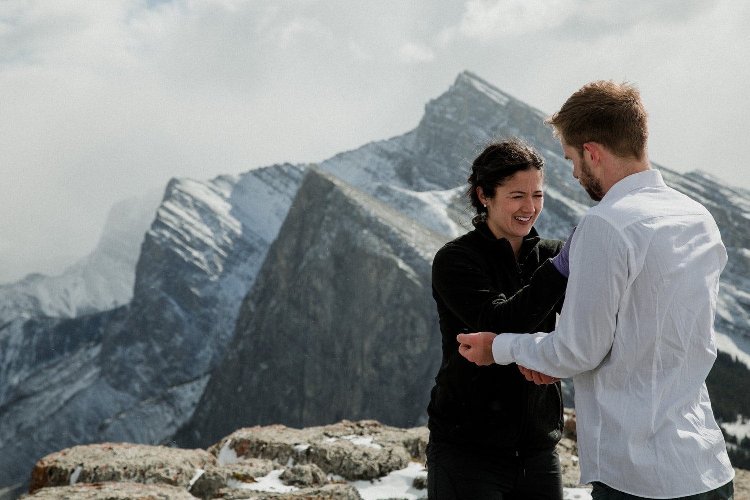 A woman help button a man's shirt on top of a mountain with views of Rundle Mountain in Banff in the background