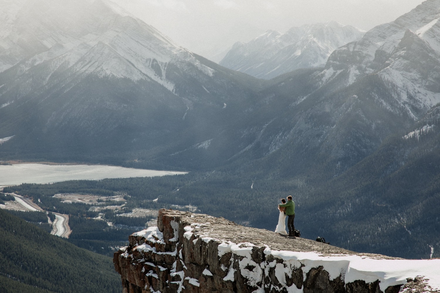 A couple stands atop a mountain and the man helps his wife into her wedding dress with epic views of mountains in the background