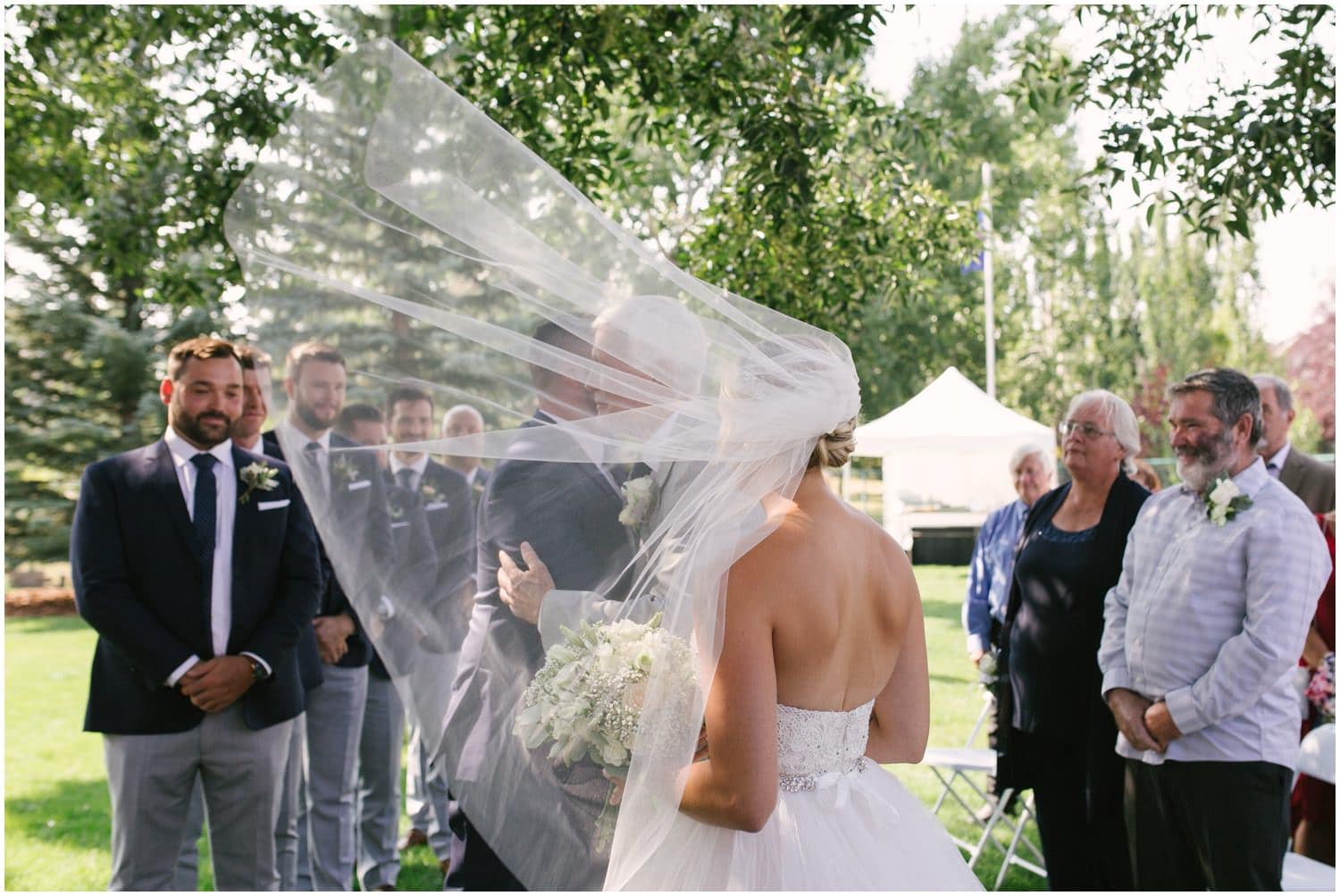 A bride's veil blows in the wind as she reaches her fiancee at the top of the aisle at their backyard wedding in Calgary