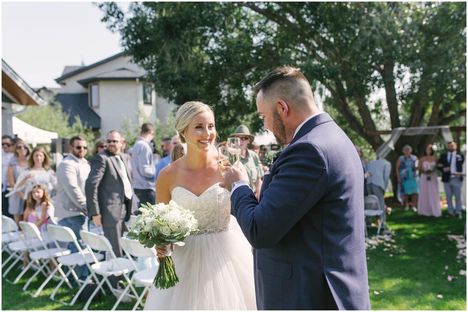 A couple cheers with champagne after completing their wedding ceremony in their backyard with guests looking on