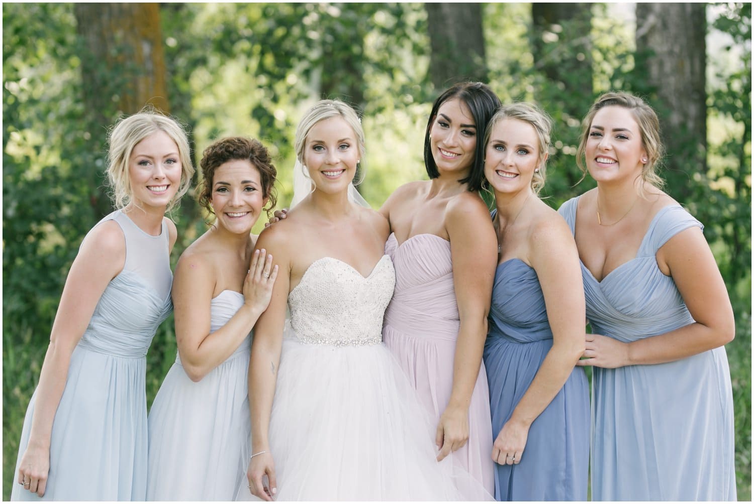 A bride and her bridesmaids in multiple shades of blue stand in front of beautiful summer green trees