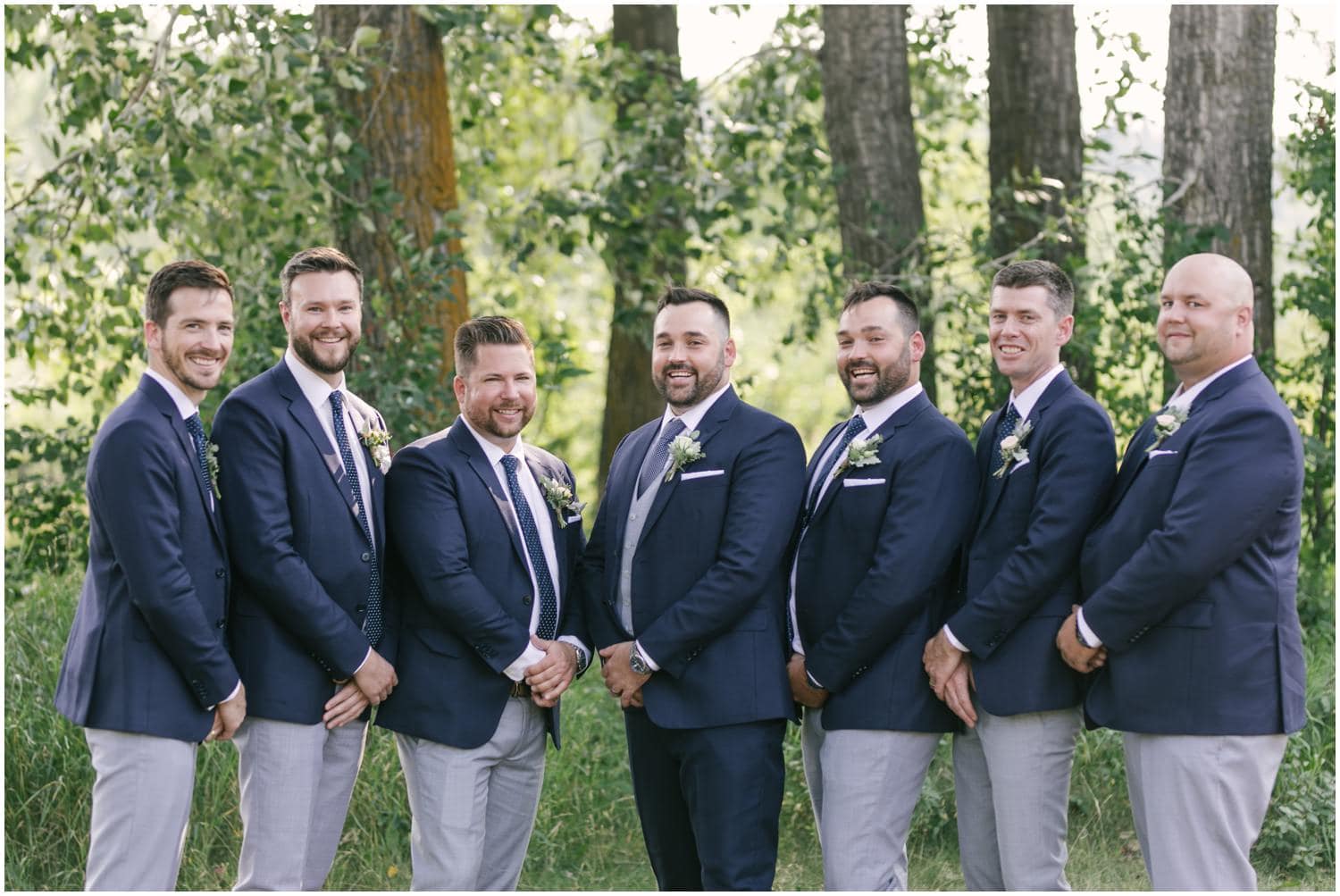 A groom and his groomsmen in navy blue suits stand in front of beautiful summer green trees