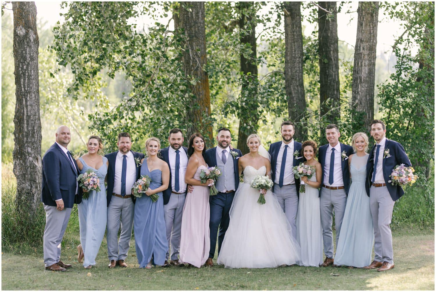 A bridal party in multiple shades of blue stand in front of beautiful summer green trees in Fish Creek Park in Calgary, Alberta