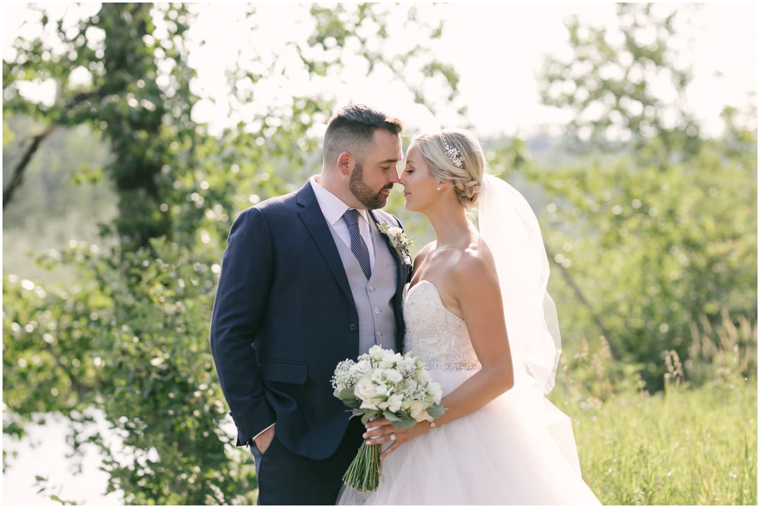 A bride and groom standing in a treed park lean in for a kiss in Fish Creek Park