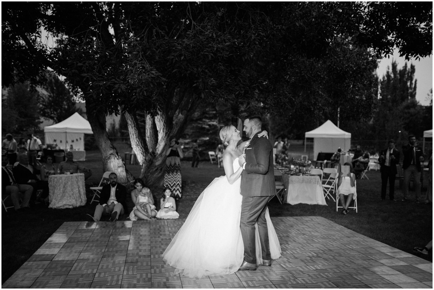 A black and white photo of a bride and groom dancing under some lights at their backyard wedding