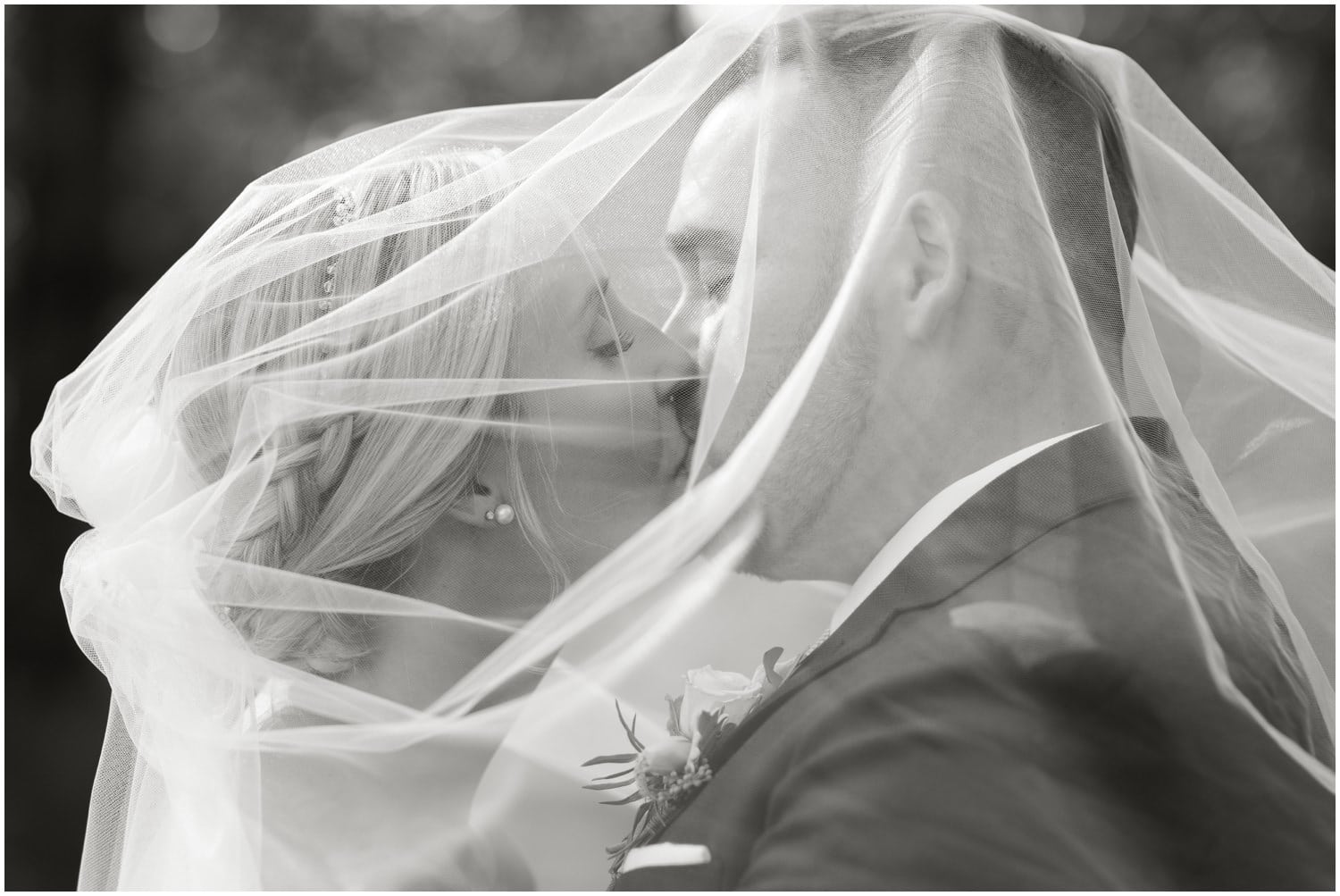 A black and white photo of a bride and groom draped under her veil sharing a kiss