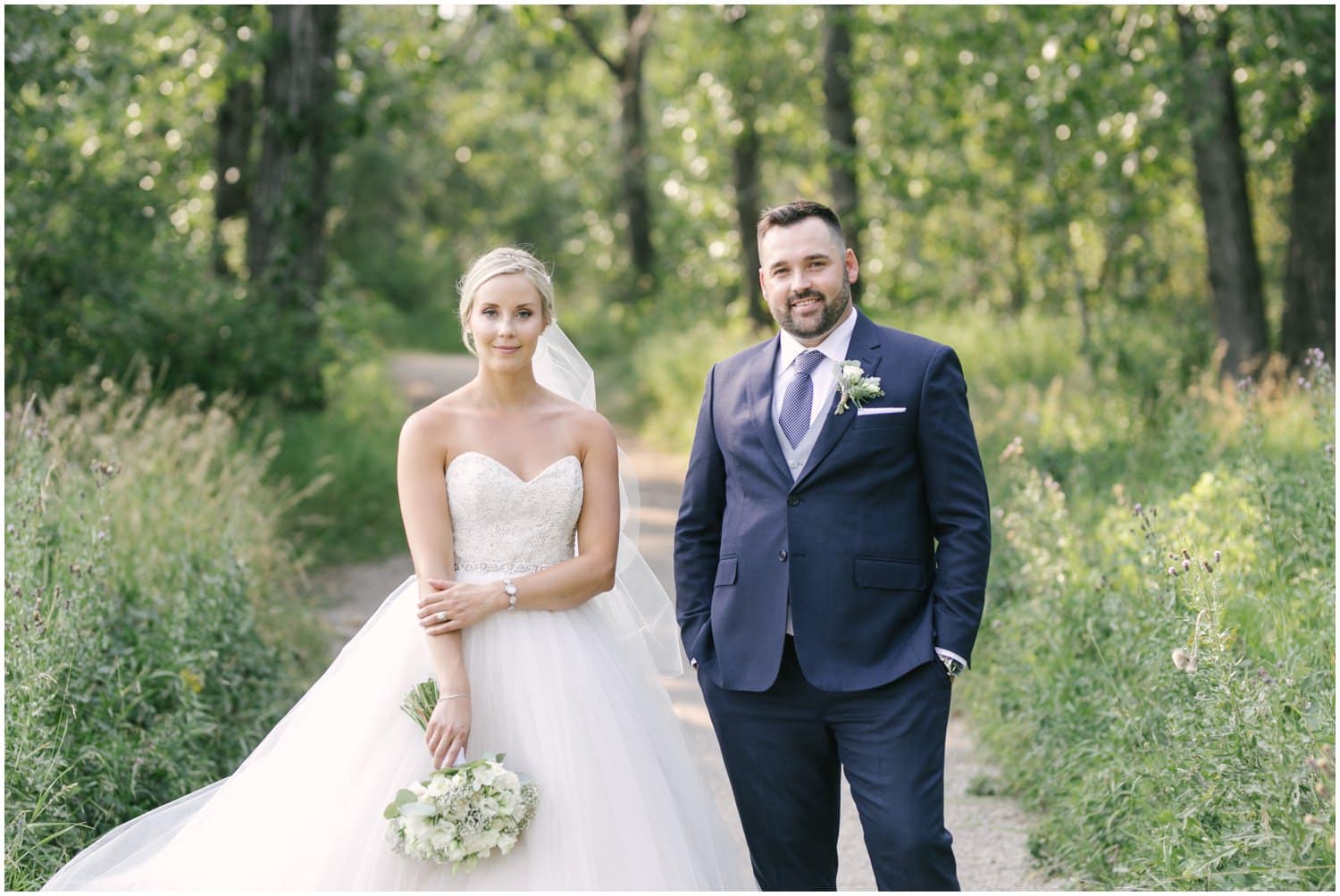 A bride and groom pose on a pathway in front of beautiful summer green trees in Fish Creek Park in Calgary, Alberta