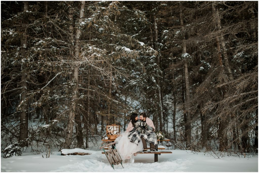 A wedding couple dressed in warm winter clothing cozy's up with a picnic in the middle of a forest