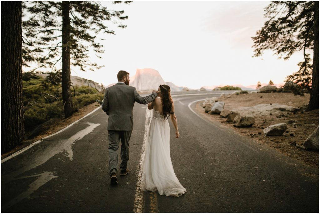 A bride kisses her grooms hand as they walk hand in hand along a road in Yosemite National Park