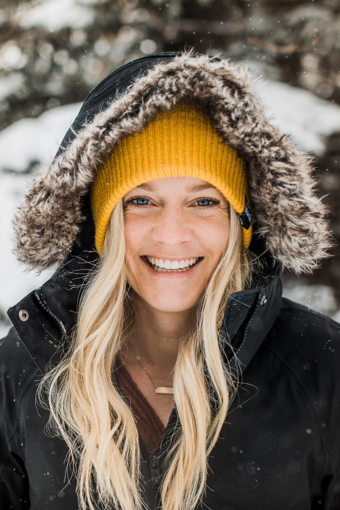 Banff elopement photographer in a bright yellow toque and black jacket smiles at the camera as snow falls around her
