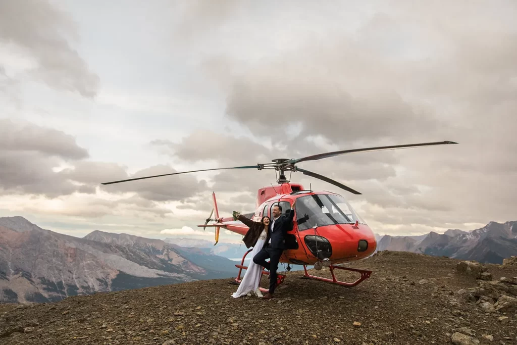 A bride and groom cheers in front of a helicopter on top of a mountain during their adventure elopement in Banff National Park