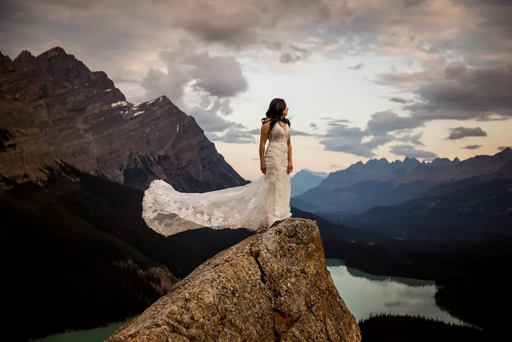 A woman in a wedding dress stand on a rock at Peyto lake for her adventure elopement