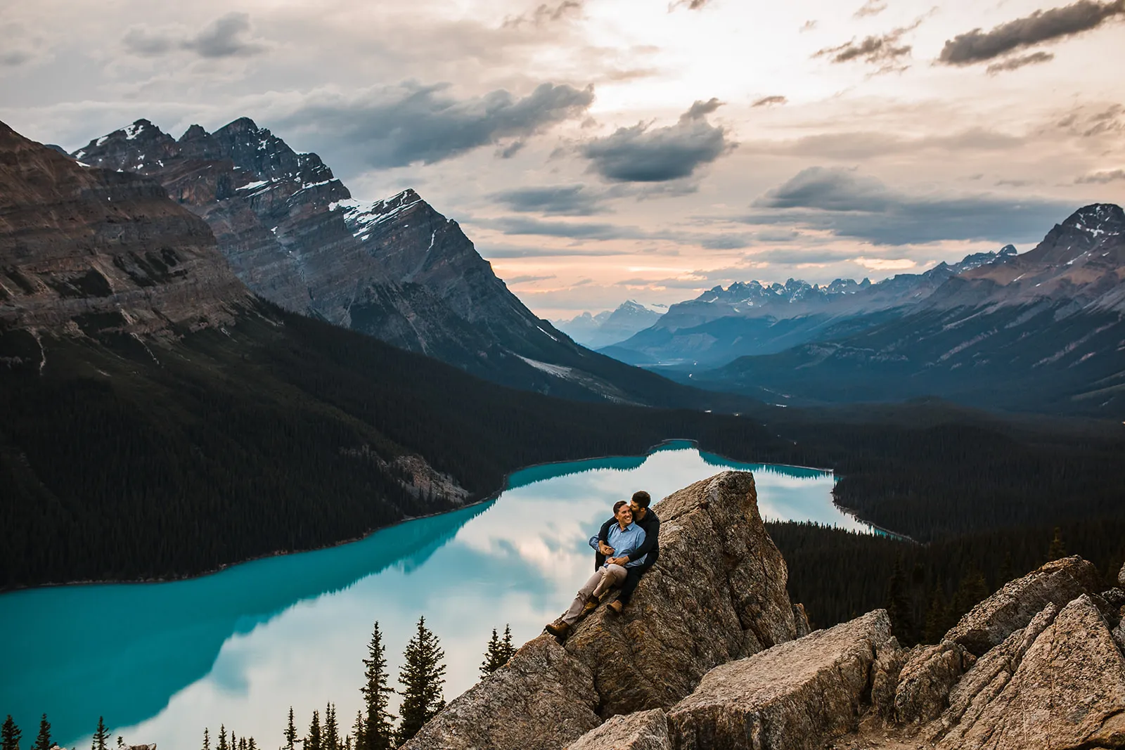 A same sex couple sit on a rock smiling at each other overlooking Peyto Lake during their same sex engagement session