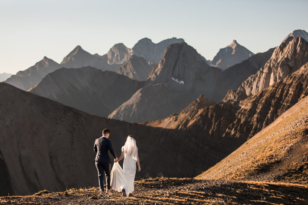 a same sex couple walks towards the mountain peaks during their sunrise hiking elopement on tent ridge kananaskis